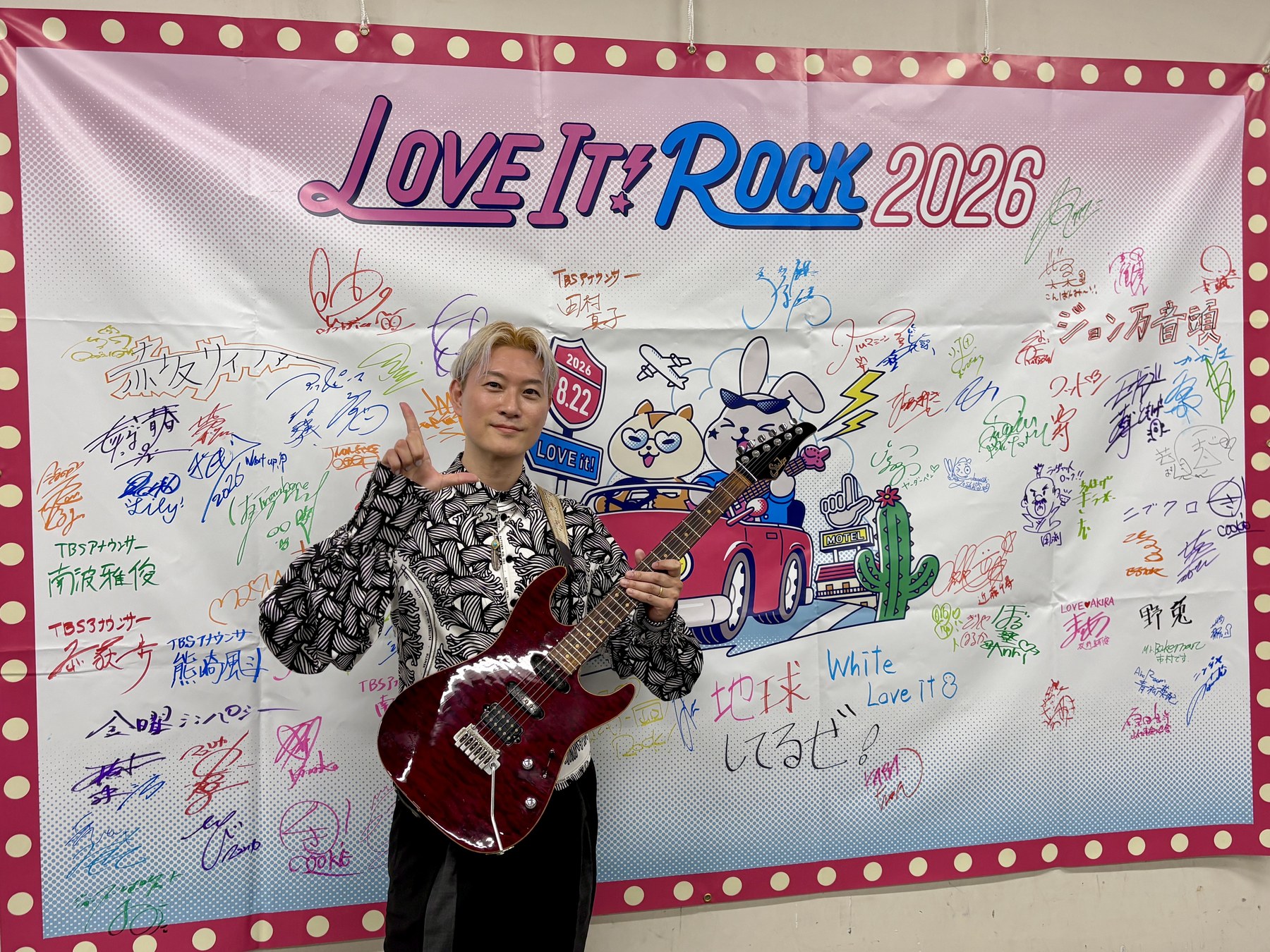LOVE IT! ROCK 2026のバナー前でギターを持つKaz